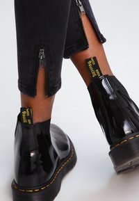 Bottines noires brillantes avec des surpiqûres jaunes portées avec un pantalon noir présentant des fermetures éclair ouvertes à l'ourlet arrière, laissant apparaître la peau.