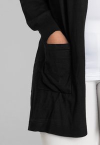 Schwarzer Cardigan mit gerippten Bündchen und Saum, ausgestattet mit einer Vordertasche. Der Stoff wirkt weich und dehnbar. Getragen über einem weißen Oberteil und hellen Hosen.