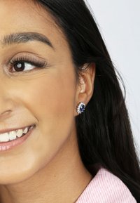 Boucles d'oreilles en pierre précieuse bleue en forme d'ovale entourées de petites pierres blanches, montées en métal argenté, portées sur l'oreille d'un modèle sur un fond clair.