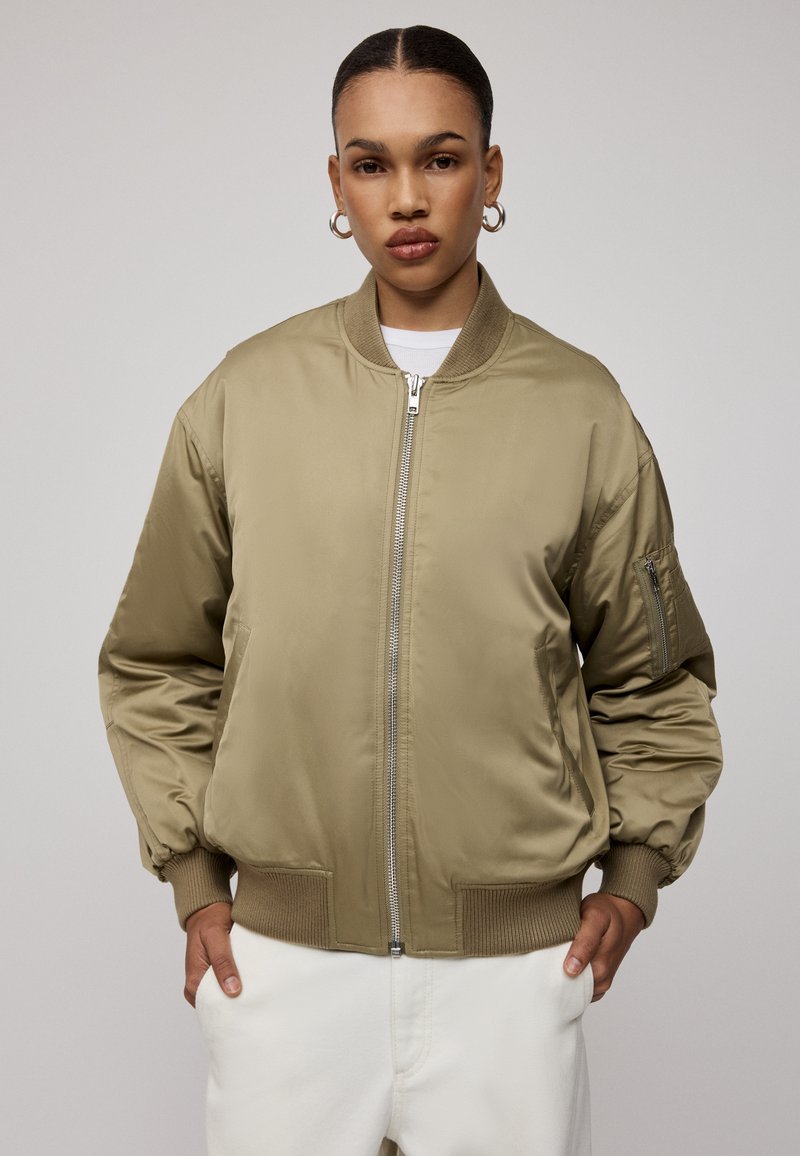 Kurtka bombowiec w kolorze khaki z gładkim, satynowym wykończeniem. Posiada zamek z przodu, ściągacze na mankietach oraz boczną kieszeń na lewym ramieniu.