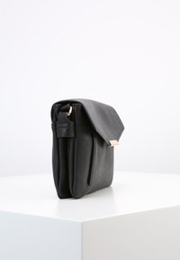 Borsa a tracolla in pelle nera con chiusura a patta e gancio dorato, in piedi su un piedistallo bianco contro uno sfondo semplice.
