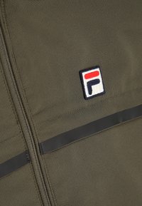 Olivgrön zip-up jacka med texturerad tyg, med ett horisontellt svart band och ett logopatch i röd, vit och blå på vänster bröst.