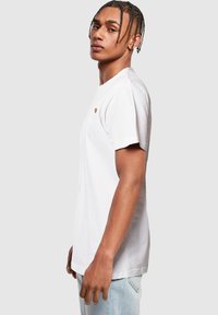 White cotton t-shirt featuring a small multicolored heart logo on the left chest, short sleeves, and a casual fit. Worn with light blue jeans.