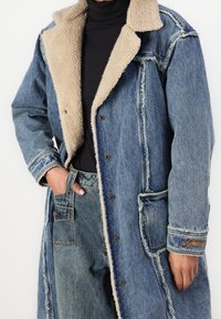 Manteau en denim avec un col en fausse fourrure beige, manches longues, texture usée, fermetures à boutons-pression et poches avant. Détail de marque brodé sur la manche.