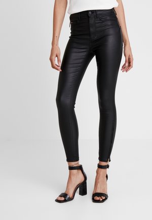 Kvinde iført højtaljede sorte skinny bukser i imiteret læder med lynlåse ved anklerne og sorte høje hælede sandaler, stående mod en ensfarvet baggrund.