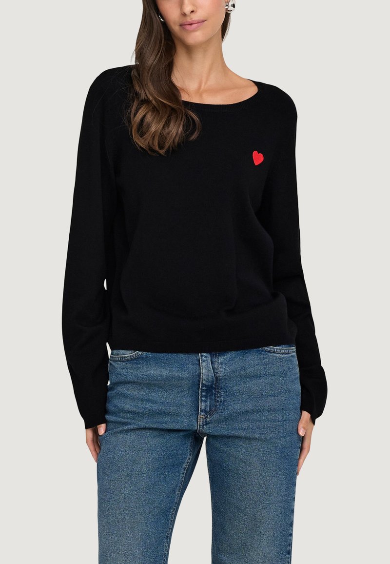 Femme portant un pull noir à manches longues avec un petit cœur rouge sur la poitrine et un jean bleu, sur un fond uni.