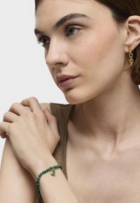 Pendientes de aro dorados con diseño floral y pulsera de cuentas verdes con un encanto en forma de corazón en la muñeca de una mujer, sobre un fondo neutro.
