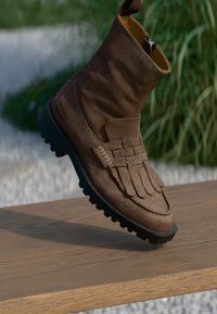 Brūni zamšādas potītes zābaki ar teksturētu virsmu, mokasīnu stila frībām, gumijas zoli un sānu rāvējslēdzēju, izlikti uz koka virsmas.