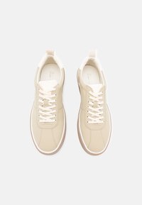 Des baskets beiges avec des accents blancs, tige en cuir lisse, bout rond, lacets plats et semelle en caoutchouc. Design minimaliste avec une esthétique moderne.
