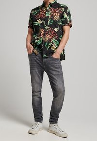 Schwarzes Kurzarmhemd mit floralen Mustern in Grün und Orange, kombiniert mit dunklen Jeans und weißen Sneakers, vor einem neutralen Hintergrund stehend.