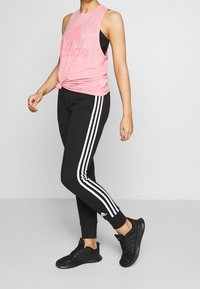 Růžová bezrukávová tank top s uzlem vpředu; černé zúžené kalhoty s bílými postranními pruhy; černé sportovní boty.