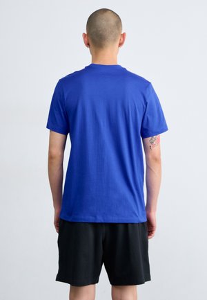 Osoba o krótkich włosach, ubrana w prosty niebieski t-shirt i czarne szorty, zwrócona tyłem, z widocznym małym tatuażem na lewej ręce.