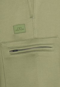 Detailní záběr na olivově zelenou látku s horizontální kapsou na zip a malou vyšitou nášivkou loga nad ní.