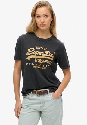 Vrouw met blond haar, gekleed in een zwart T-shirt met het vintage "SuperDry"-logo en lichtblauwe jeans met een bruine riem, poserend met één hand in haar zak.