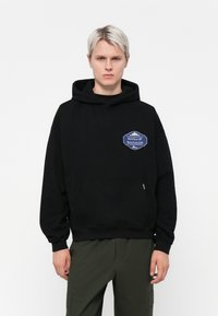 Zwarte hoodie van katoen, met een voorzak en een blauw geborduurd label met "Notes of Nostalgia" op de linkerkant van de borst.