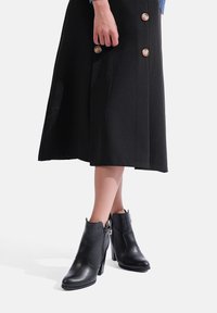 Stivaletti neri in pelle con dettaglio a zip, tacco alto e punta tondeggiante, abbinati a una gonna nera con grandi bottoni.