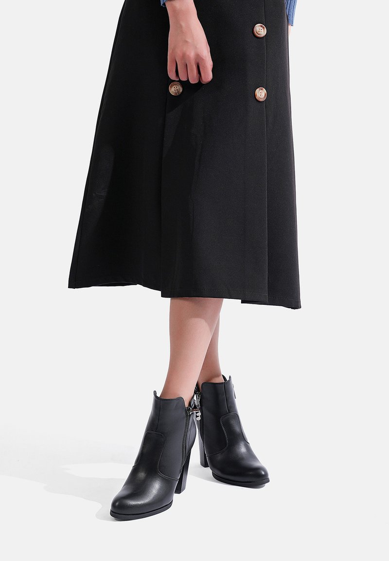 Stivaletti neri in pelle con dettaglio a zip, tacco alto e punta tondeggiante, abbinati a una gonna nera con grandi bottoni.