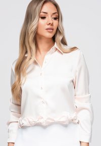 Femme aux longs cheveux blonds portant une chemise rose clair boutonnée avec les manches retroussées, regardant légèrement vers le bas et sur le côté.