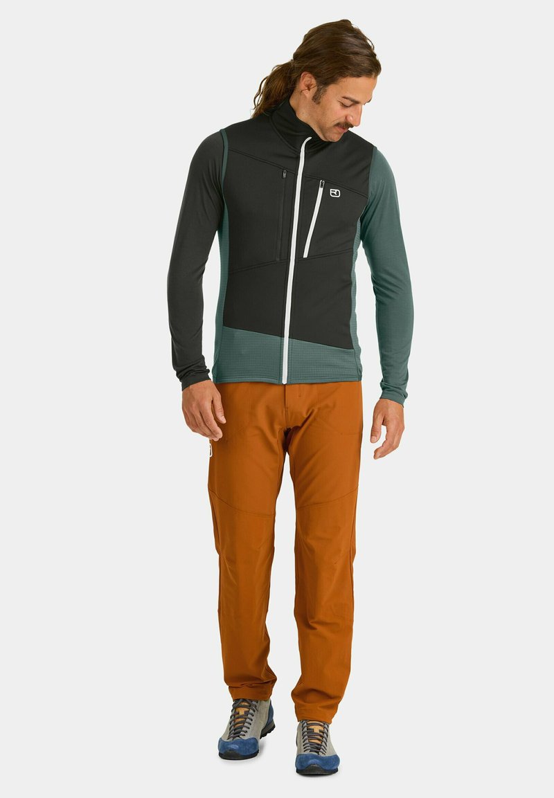 Schwarwest und türkisfarbener Reißverschluss-Weste mit zwei Vorderstaschen, kombiniert mit orangenen Kletterhosen und grauen Wanderschuhen mit blauen Akzenten.