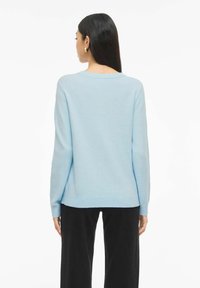Femme aux longs cheveux foncés portant des boucles d'oreilles en or, un pull à manches longues bleu clair et un pantalon noir, vue de dos sur un fond blanc.