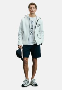 Weißer Reißverschluss-Hoodie mit schwarzen Akzenten, T-Shirt mit Grafikdruck, navyfarbene Shorts und graue Sneaker; hält eine schwarze Mütze in einer Hand.