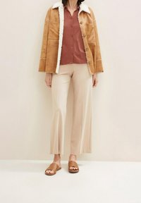 Frau trägt eine hellbraune Lammfelljacke über einem rostfarbenen Hemd mit Knöpfen, cremefarbene Hosen mit weitem Bein und braune offene Sandalen mit Riemen vor einfarbigem Hintergrund.