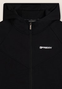 Sudadera negra con cremallera, fabricada en un material suave, que cuenta con una capucha forrada de malla, costuras en ángulo y un logo blanco en el pecho.