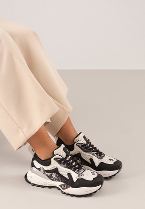 Zapatillas en blanco y negro con detalles de malla y texturizados, que presentan una suela gruesa y cordones. Combinadas con pantalones anchos beige.