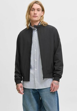 Black bomber jacket with a smooth texture, zip closure, and ribbed cuffs, worn over a blue and white striped shirt with buttons.