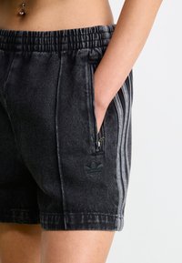 Calções de ganga preta com cintura elástica, apresentando bolsos laterais e detalhes decorativos de três riscas. Logotipo verde escuro no bolso.