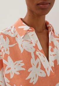 Korte mouwen, koraalkleurige blouse met een wit bloemenpatroon. Heeft een kraag, V-hals en lichtgewicht stof, wat een ontspannen pasvorm benadrukt.