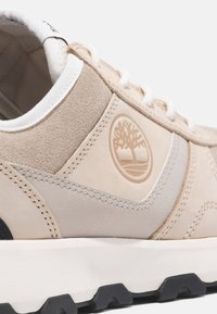 Sneaker in suede e pelle beige con superficie testurizzata, dettagli cuciti, lacci bianchi e una patch con logo. Suola in gomma con battistrada.