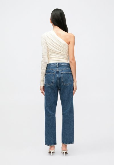 Top de um ombro só em off-white com franzidos, combinado com jeans azul escuro de perna reta. O top apresenta tecido texturizado; os jeans têm bolsos clássicos.