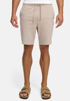 Mężczyzna w beżowych szortach z sznurkiem i dopasowanych sandałach na płaski obcas stoi na białym tle.