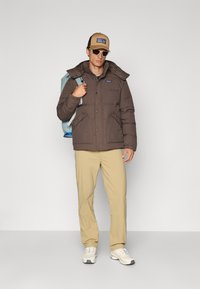 Brun isoleret pufferjakke med aftagelig hætte, beige bukser og hvide sneakers. Accessories inkluderer en tan cap og solbriller.