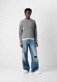 Mann im grauen Strickpullover, hellblauen, rissigen, weit geschnittenen Jeans und weißen Sneakers, der vor einem schlichten weißen Hintergrund steht.