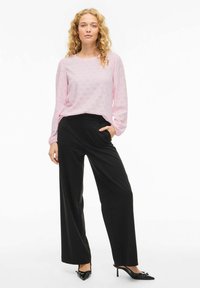 Woman with curly blonde hair wearing light pink long-sleeve blouse and high-waisted wide-leg black trousers with black heels, standing.