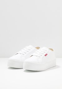 Baskets en toile blanche à semelle épaisse, dotées d'un bout rond, d'un système de laçage et d'un petit logo rouge sur le côté.