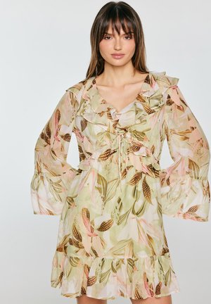 Kobieta ubrana w przejrzystą, kwiecistą sukienkę z długim rękawem, z falbankami przy kołnierzu, rękawach i dole, stojąca na jednolitym jasnym tle.