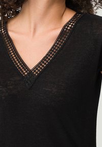 Svart V-ringad topp med en texturerad spetskant runt halslinningen. Mjuk, lätt tyg med en något genomskinlig look.