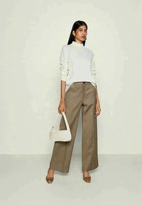 Balta megzta palaidinė su aukštu kaklu, laisvos rudos kelnės ir balta rankinė. Rudi bateliai su raštais puošia aprangą. Modelis stovi priešais šviesią foną.