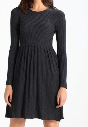 Femme portant une robe en maille côtelée gris foncé à manches longues avec une jupe plissée, debout devant un fond blanc.