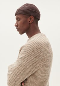 Caciula tricotată maro cu o textură ribbed, asociată cu un pulover gros din tricot bej cu guler rotund.