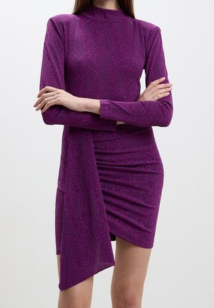 Frau trägt ein langärmeliges, violettes, strukturiertes Kleid mit hohem Kragen und asymmetrischem Saum, die Arme vor dem Oberkörper verschränkt.
