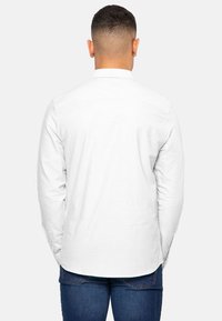 Camisa blanca de manga larga con cuello abotonado, textura suave y dobladillo recto. La vista trasera muestra un diseño sencillo y un ajuste entallado.