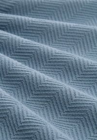 Helles blaues Gewebe mit einem Fischgrätenmuster, das eine strukturierte, weiche Optik und Webdetails aufweist, sanft in Falten drapiert.