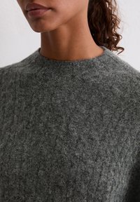 Grauer Strickpullover mit rundem Ausschnitt, strukturiertem Stoff und weicher Optik, der subtile Abstufungen in der Schattierung und ein gemütliches Design aufweist.