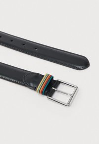 Cinturón de cuero negro con hebilla de metal plateado, que presenta un detalle de tela a rayas en rojo, verde y amarillo cerca de la hebilla.