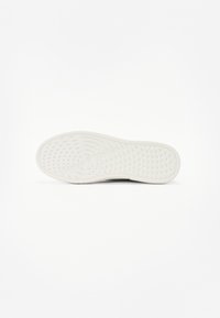 Podešev bílé tenisky s kruhovými a trojúhelníkovými gripovými vzory, na které je uprostřed vyraženo jméno značky.