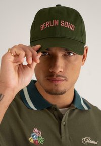 Căciulă verde de baseball cu broderie roz "BERLIN SOHO", purtată într-un unghi. O cămașă polo verde prezintă un detaliu de broderie florală multicolor.